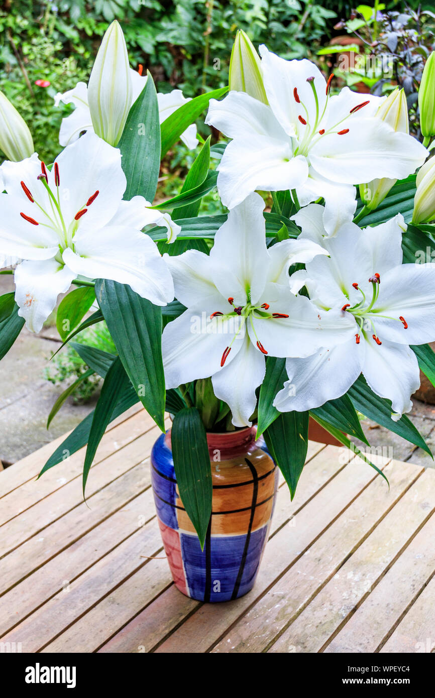 a bunch of white lilies arranged in a colourful vase on a wooden garden table, London, UK Stock Photo