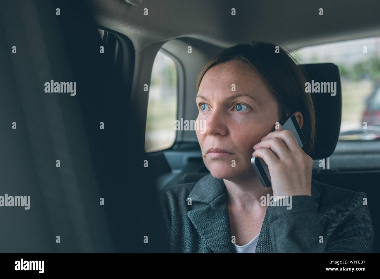 Doubtful businesswoman talking on mobile phone on car back seat, business on the move concept. Elegant adult caucasian female person using smartphone Stock Photo