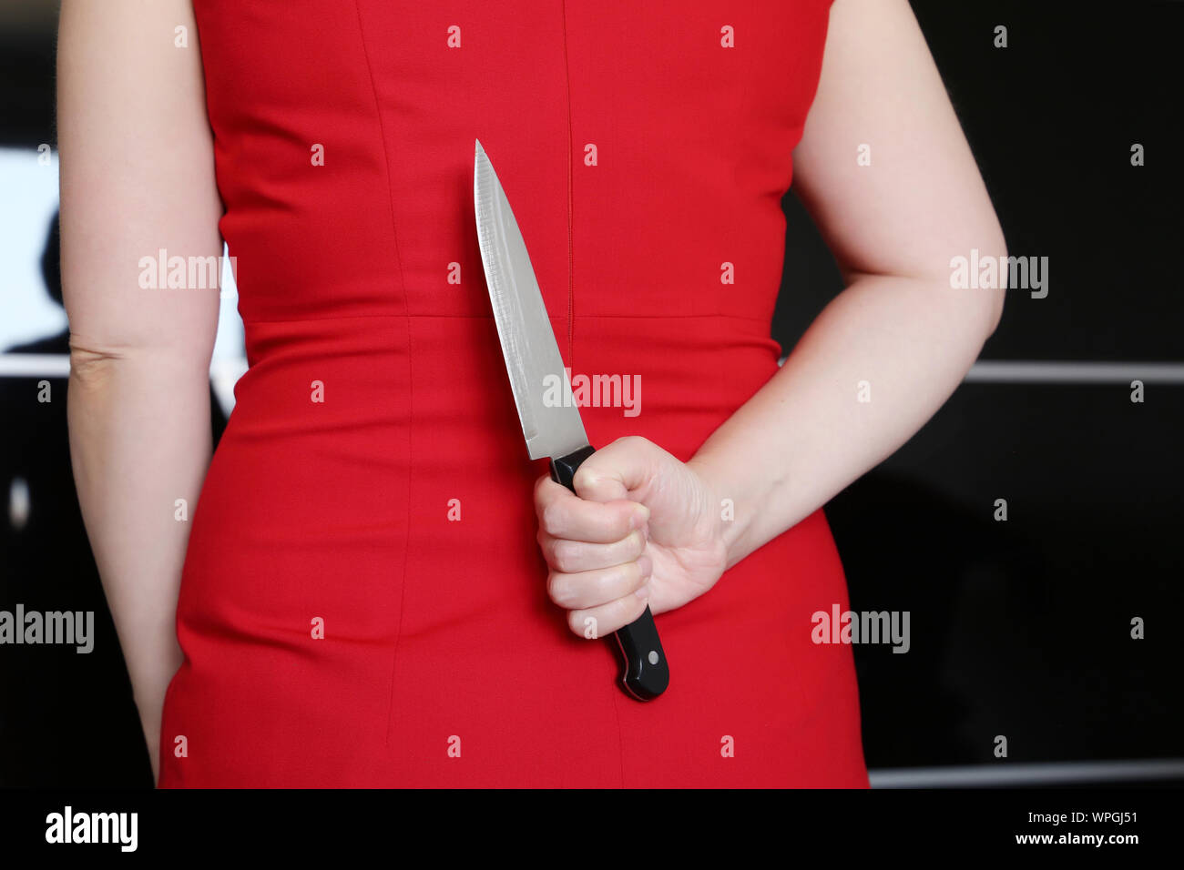 Woman in red dress standing with a knife in hand behind her back. Concept of crime, self defense, murder, maniac, revenge Stock Photo