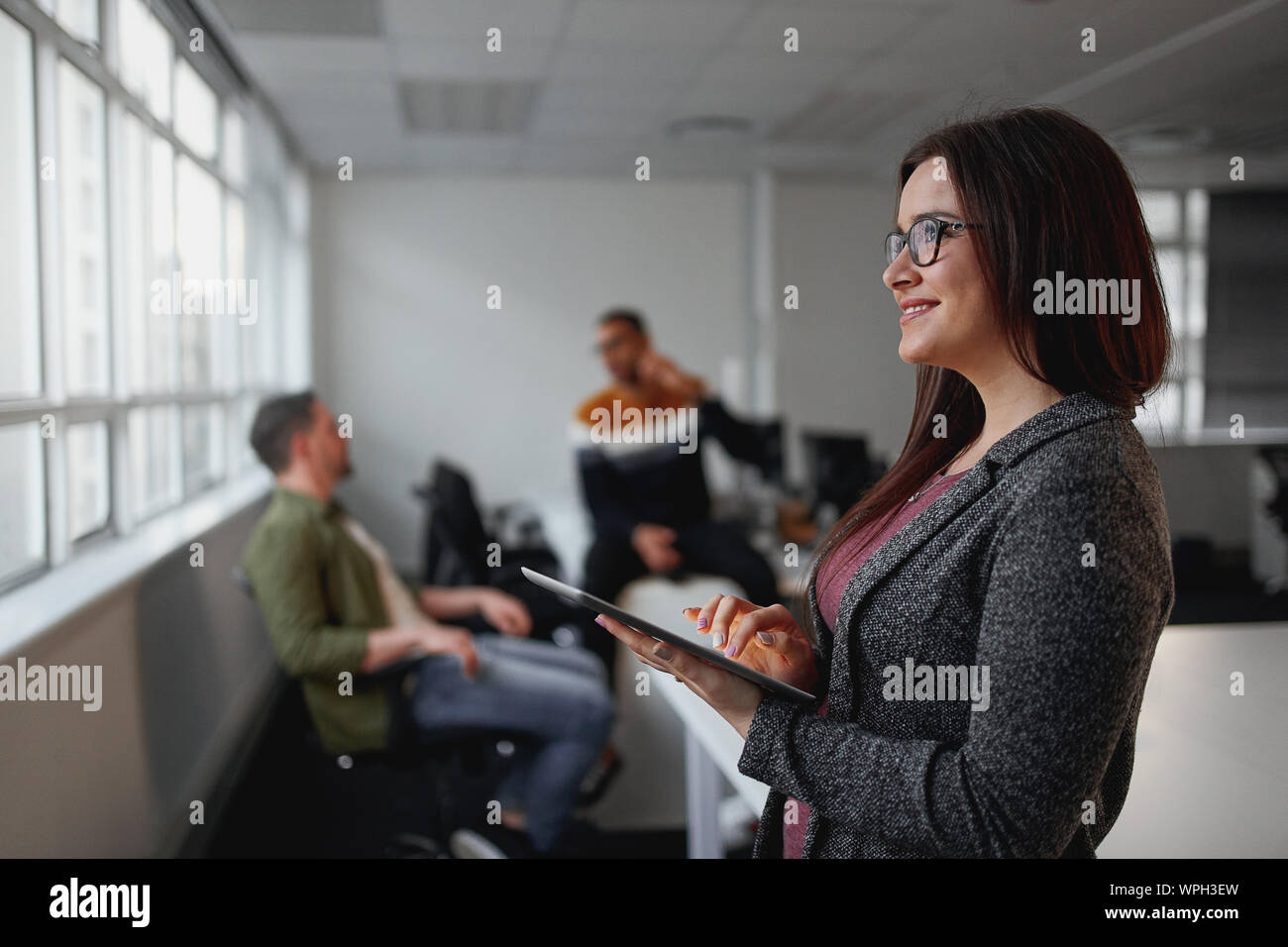 Thoughtful young businesswoman looking away while holding digital tablet in front of colleagues relaxing in office at background Stock Photo