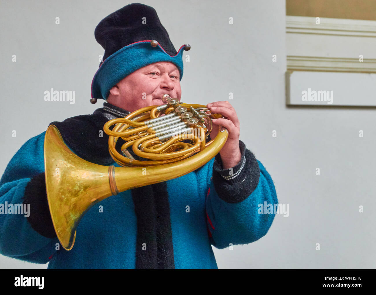 Dresden, Germany, December 14., 2018:Horn blower in a historical blue dress plays on the old horn outdoors in front of a white wall Stock Photo