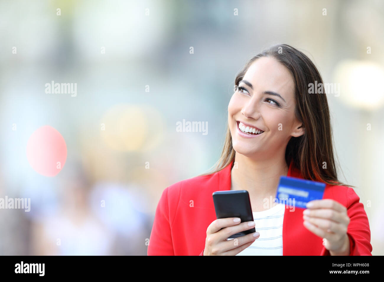 Happy woman in red holding credit card and smart phone thinking looking at side in the street Stock Photo