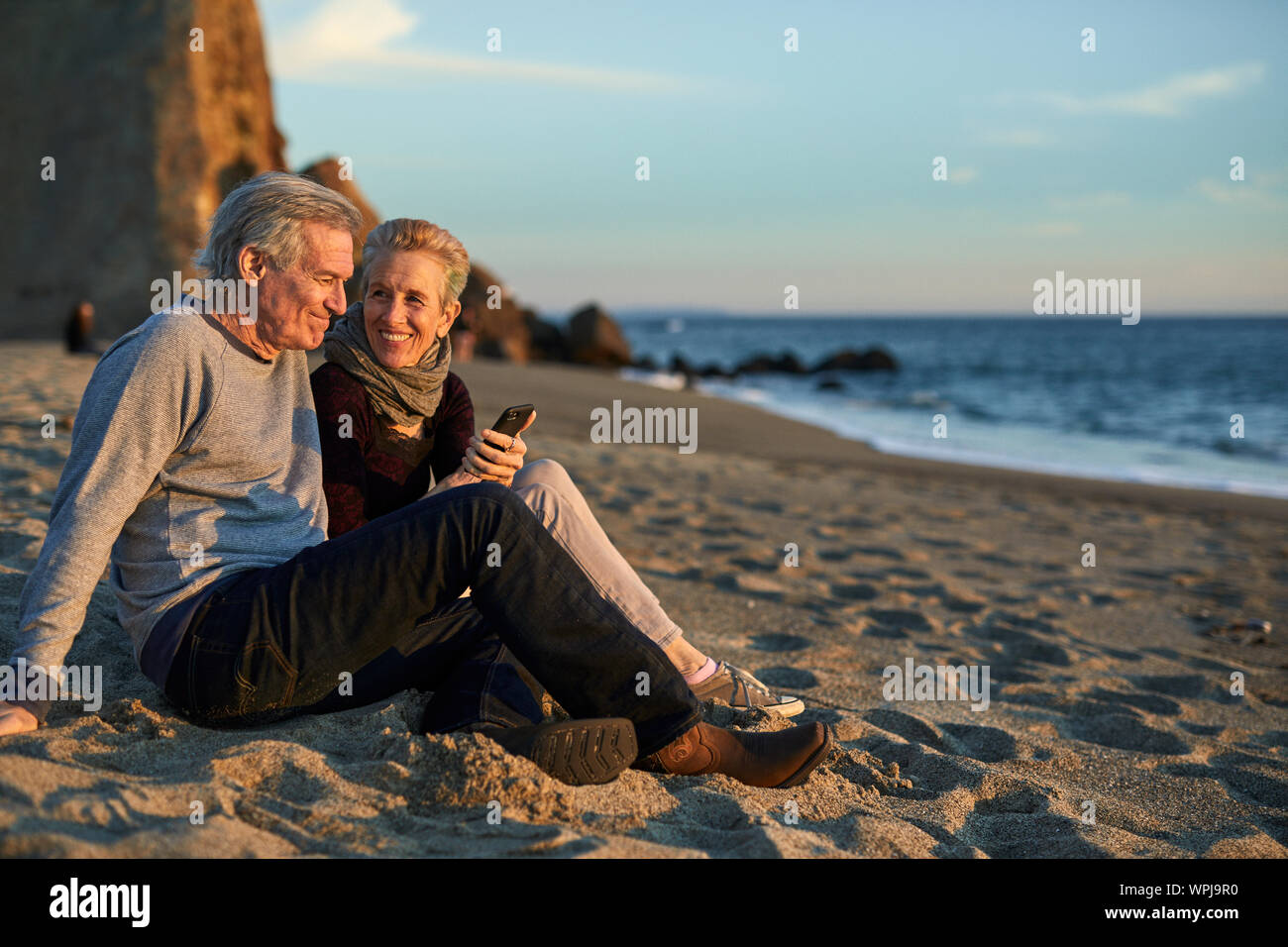 Senior couple using phone while sitting on sand at beach during sunset Stock Photo