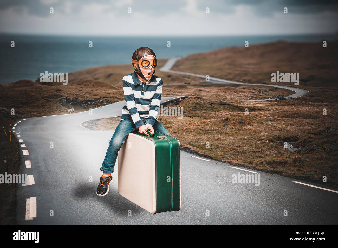child dressed as an airman on a suitcase imagines to live an adventure flying on a lost road Stock Photo