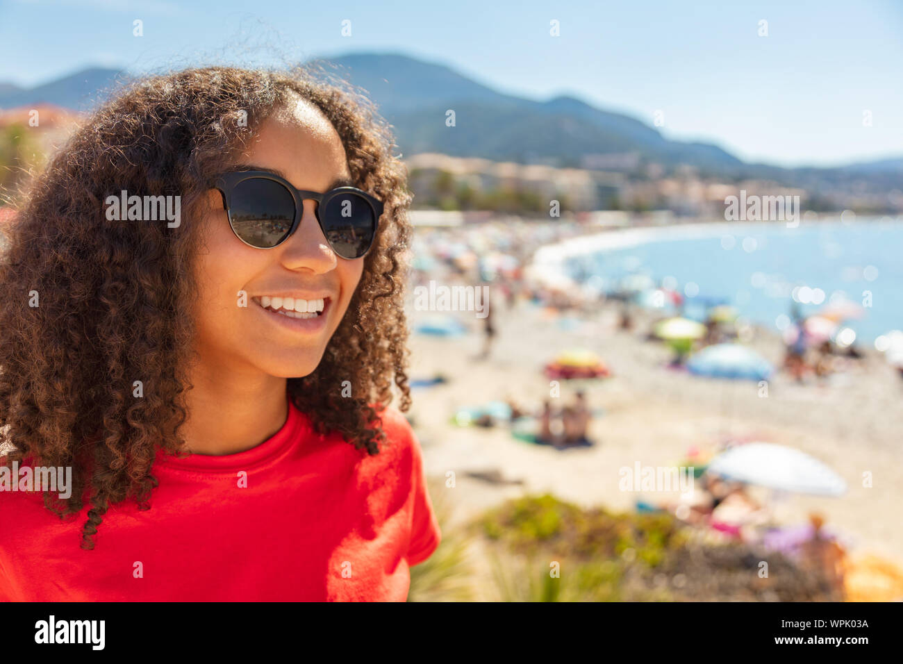 Beautiful mixed race biracial African American female girl teenager young woman on vacation with perfect teeth, wearing sunglasses on a beach. Shot in Stock Photo