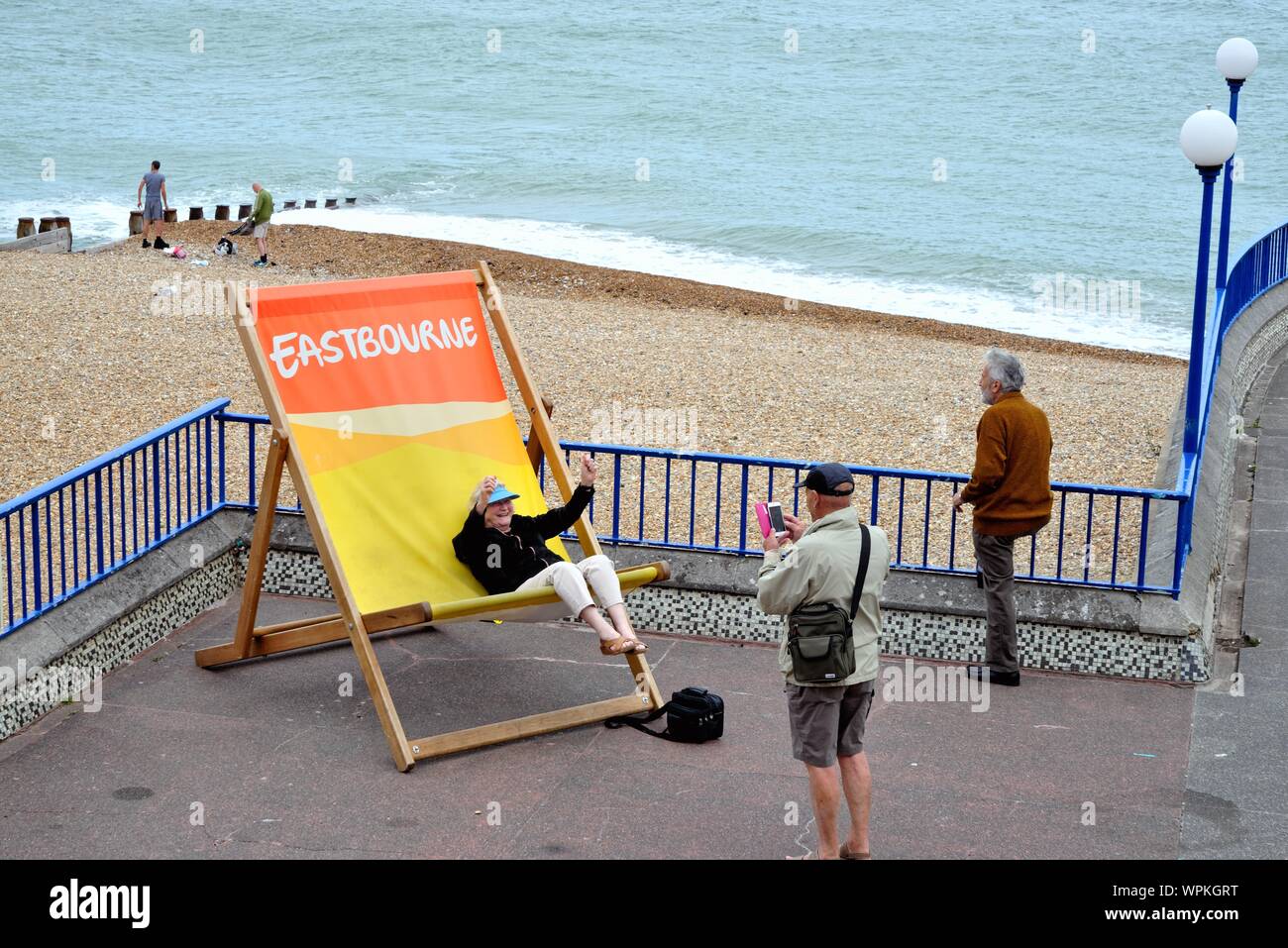 An old lady sitting on an oversized deckchair having fun and waving her arms while being photographed on Eastbourne seafront, Sussex England UK Stock Photo