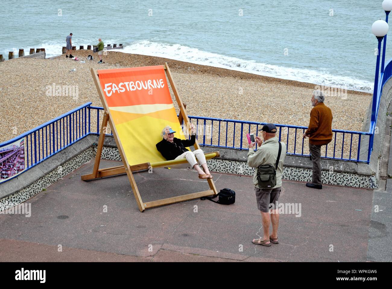 An old lady sitting on an oversized deckchair having fun and waving her arms while being photographed on Eastbourne seafront, Sussex England UK Stock Photo