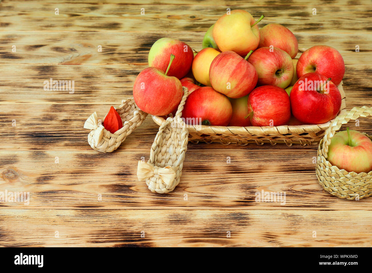 Red apples and autumn flowers lie in a wicker plate on a wooden table. Wicker shoes and apples in a basket. Healthy food and lifestyle, vegetarianism. Stock Photo