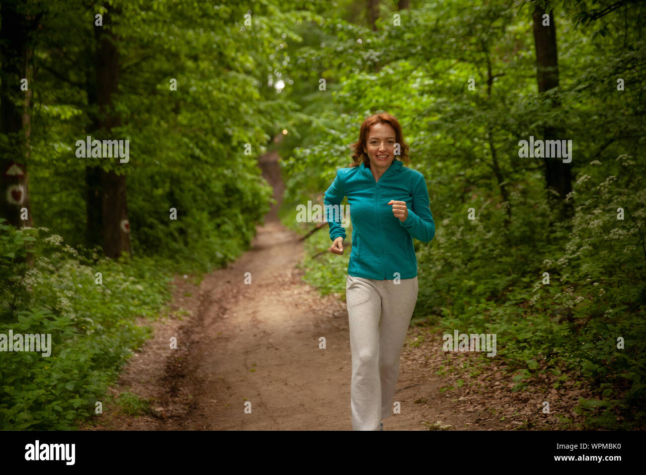 One mature woman, 40-45 years old, jogging in a forest outdoors, in nature. Wearing sport clothing in Spring. Stock Photo