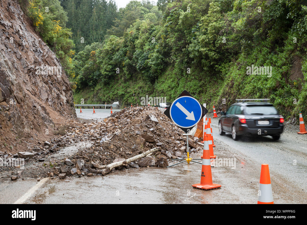 Automobile car driving past rockflall, rocks, landslide debris, traffic cones and warning signs on roadway highway. Dangerous road conditions hazards. Stock Photo