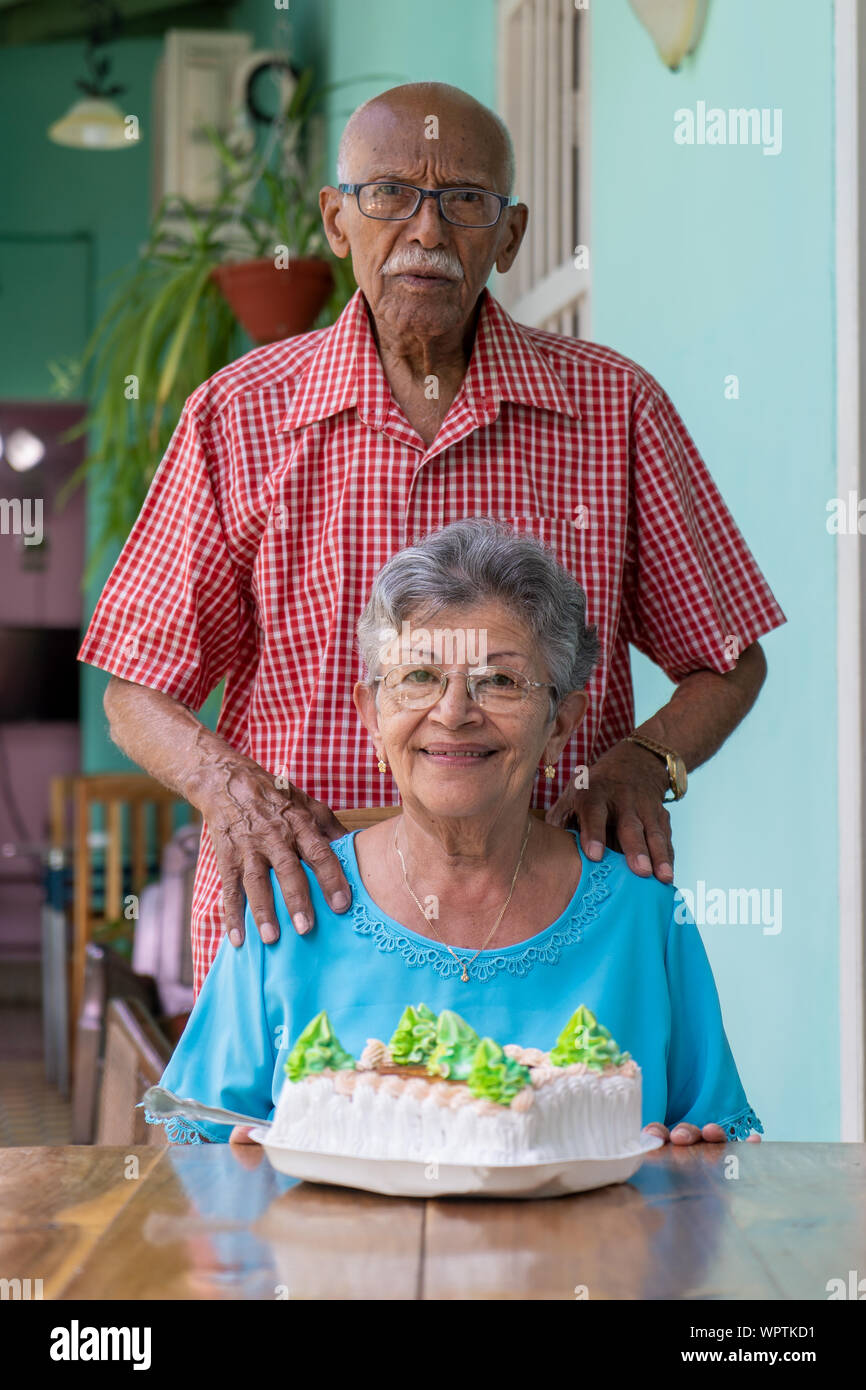 An elderly couple, the woman sitting and a man stand up and a cake on a table Stock Photo