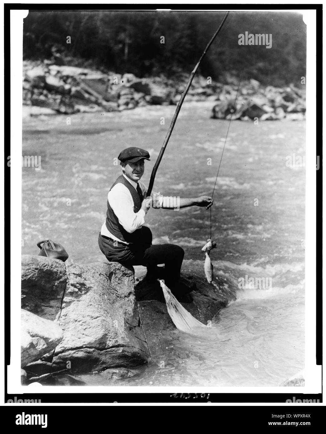 Man seated on rock, with two fish on line, by stream, possibly the River, Washington, D.C. area Stock Photo
