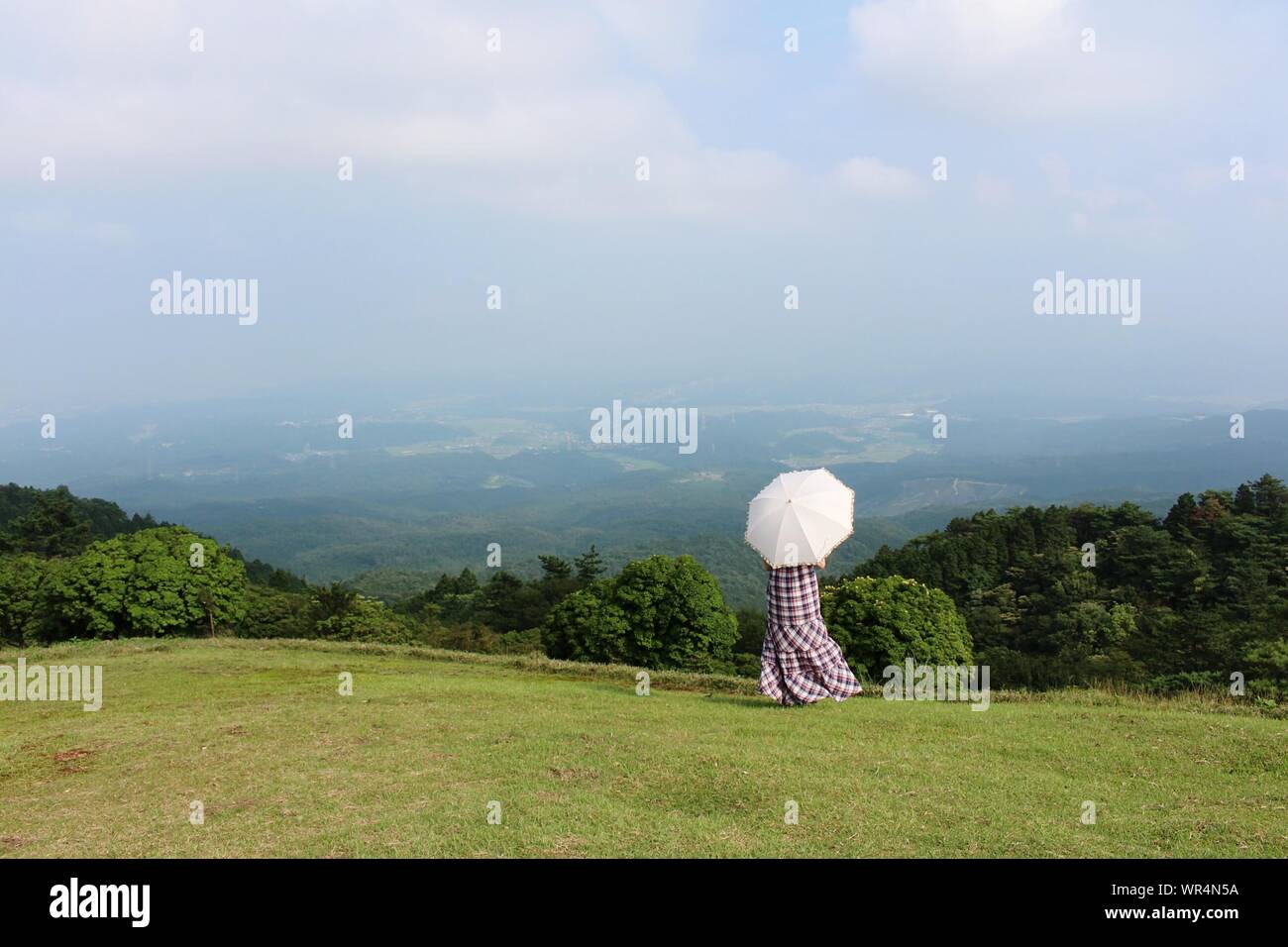 Rear View Of Woman Wearing Long Dress With Umbrella Standing On Grass Against Mountain Stock Photo