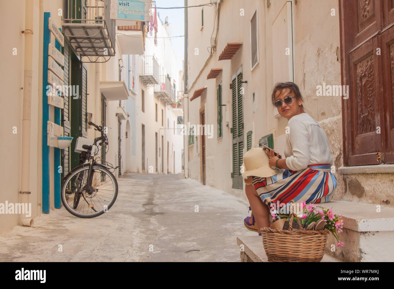 Young woman sitting on door steps in a typical alley on Ponza island town, Italy. Fashion dress and sunglasses, white building. Stock Photo