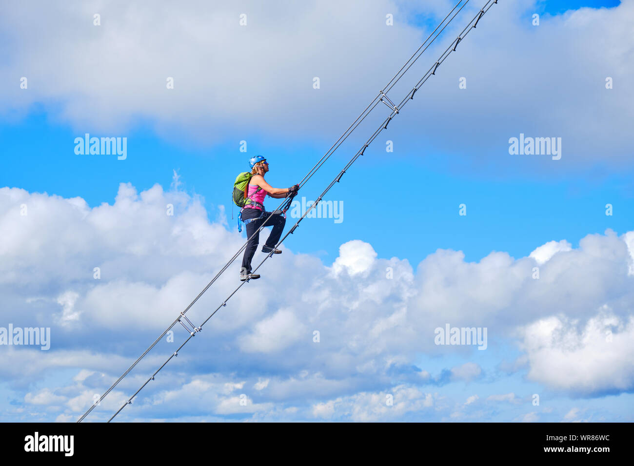 Stairway to heaven concept. Woman climbing a diagonal via ferrata ladder in Donnerkogel mountains, Austria, near Gosau, with white clouds and blue sky Stock Photo