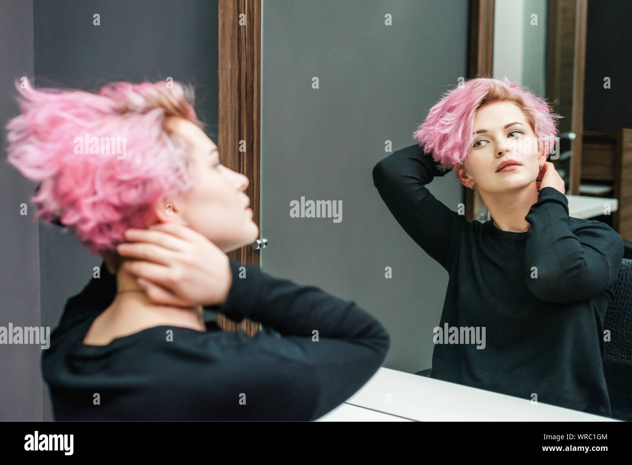 Beautiful hairstyle of young woman looking in the mirror after dying hair and making highlights in beauty salon. Close up short pink hairdo Stock Photo