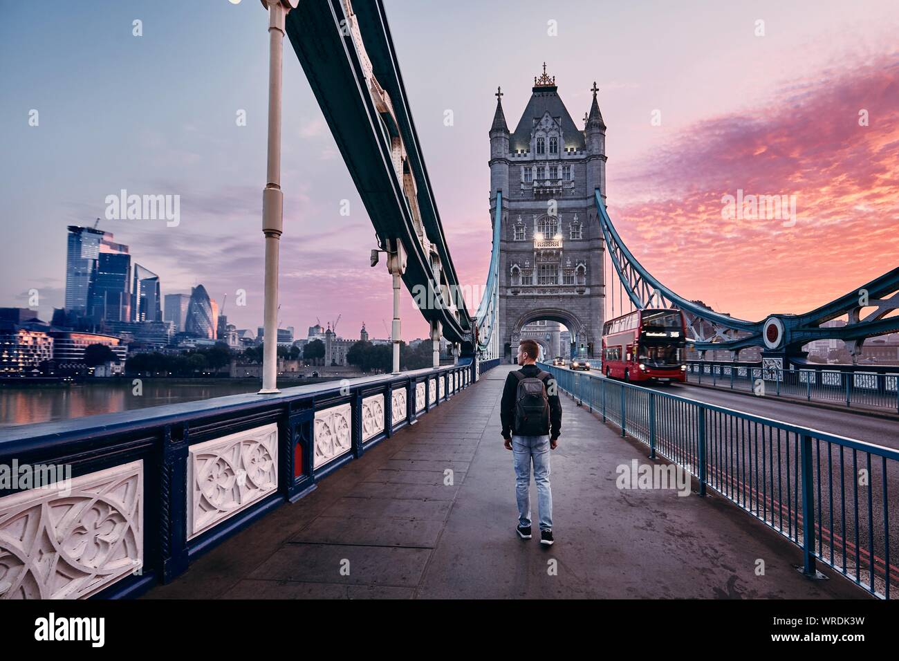 Young man with backpack walking on Tower Bridge against cityscape with skyscrapes at colorful sunrise. London, United Kingdom Stock Photo