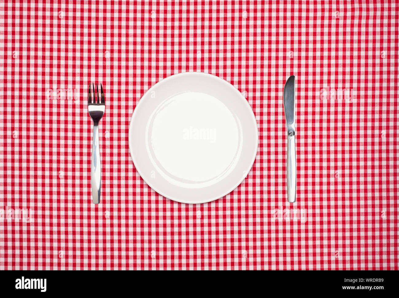 Place setting, round white plate, knife and fork, from above on a red gingham tablecloth Stock Photo