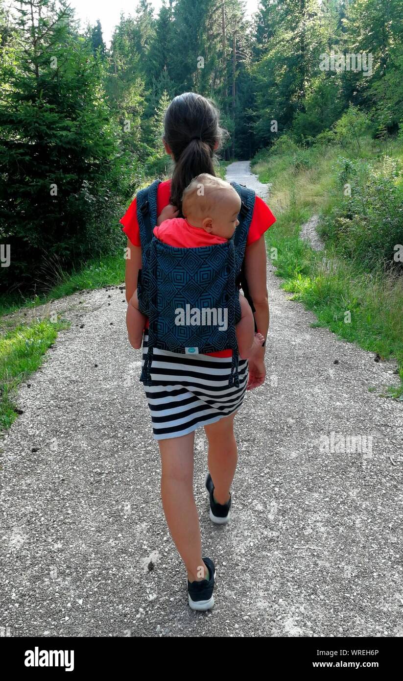 Rear View Of Woman Carrying Her Baby In Baby Carrier While Walking On Footpath Stock Photo