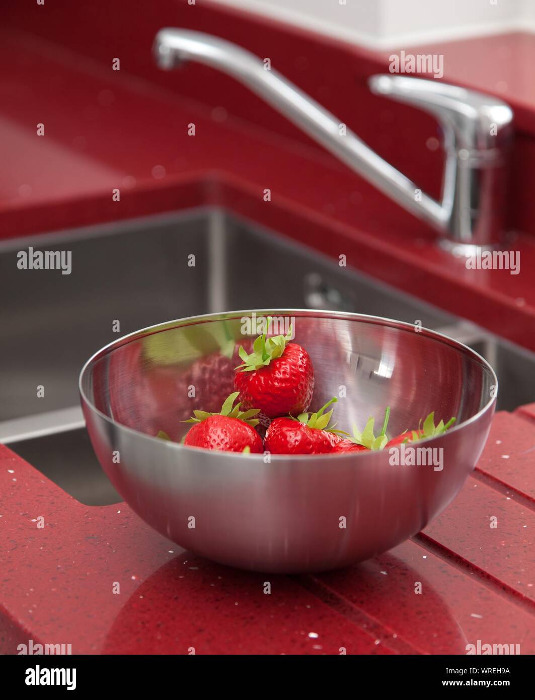 Strawberries in a stainless steel bowl beside a kitchen sink set into bright red worktop. Stock Photo