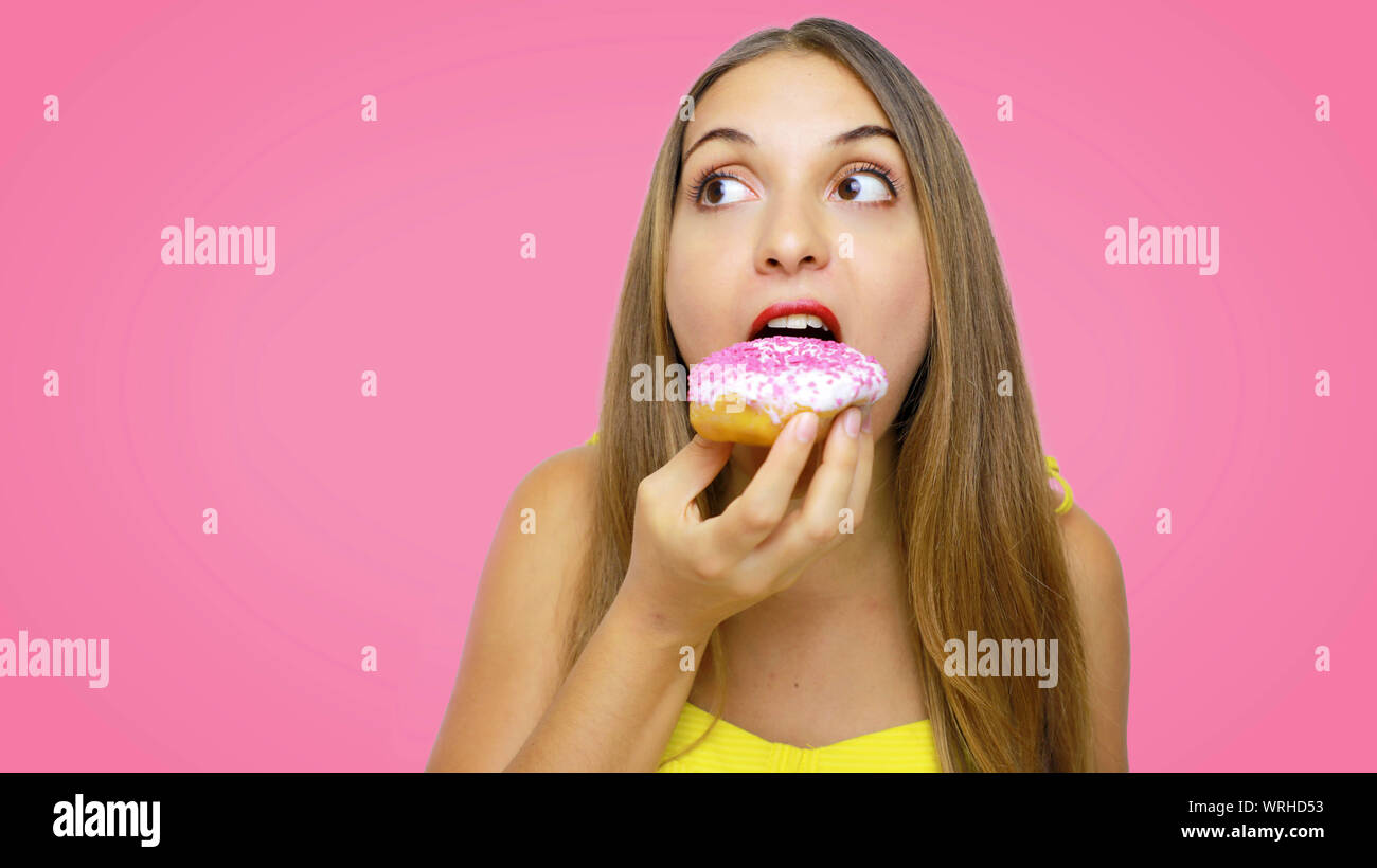 Hungry young girl eating donut over pink background looks to the side