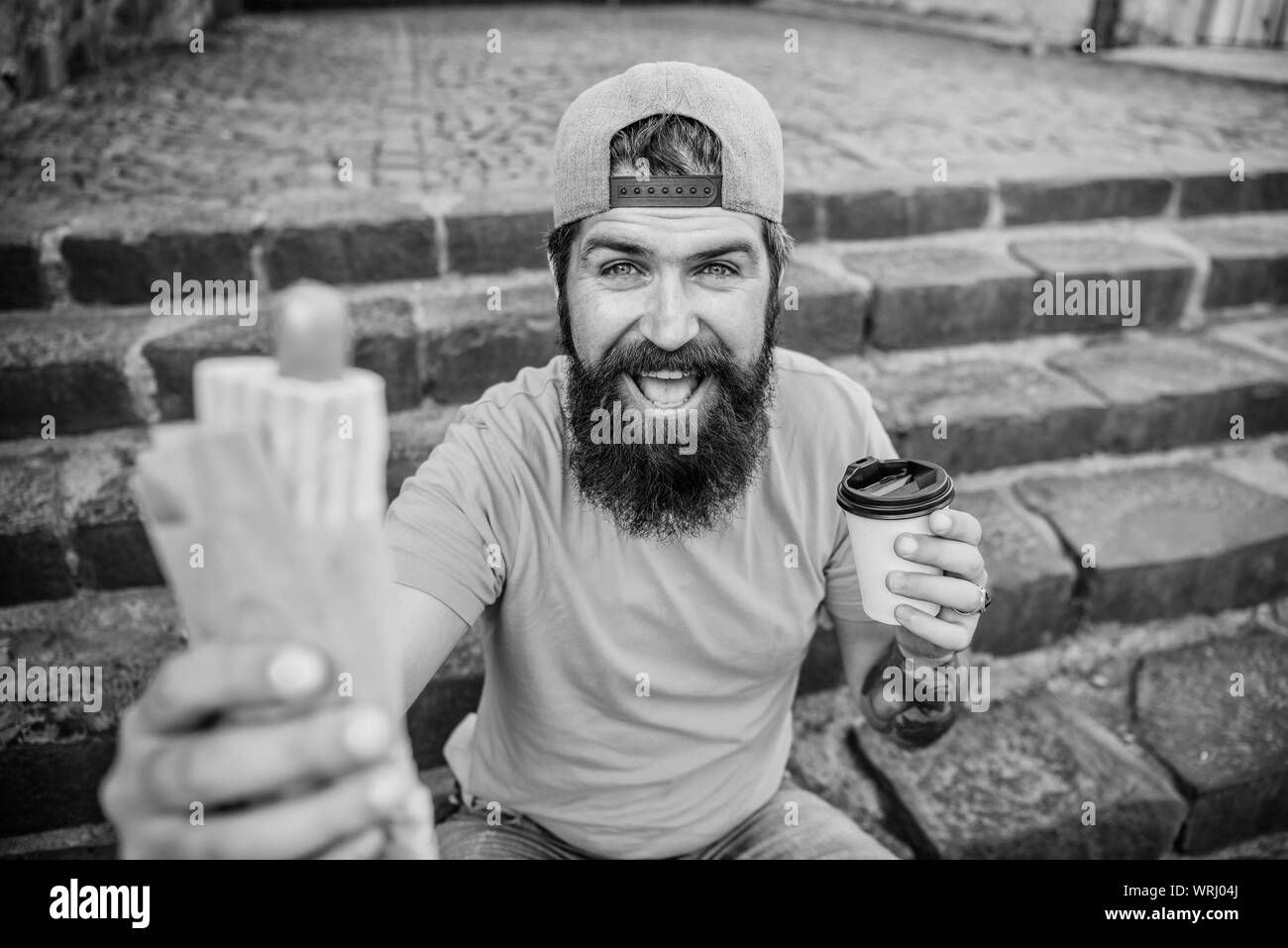 Snack for good mood. Guy eating hot dog. Man bearded eat tasty sausage and drink paper cup. Street food concept. Urban lifestyle nutrition. Junk food. Carefree hipster eat junk food while sit stairs. Stock Photo