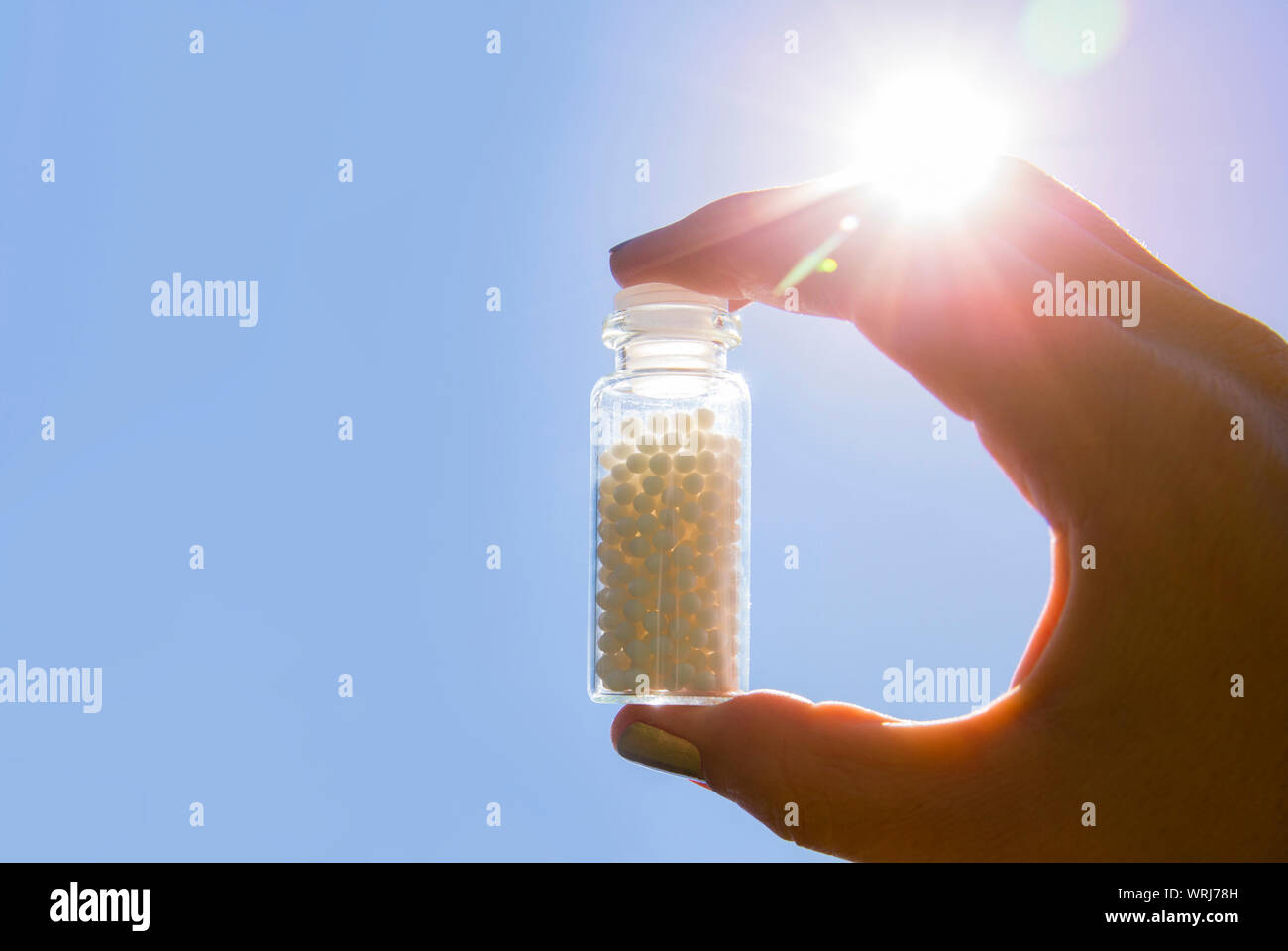 Selective focus on person hand holding glass jar full of small white round homeopathy pills against blue sky background. Stock Photo