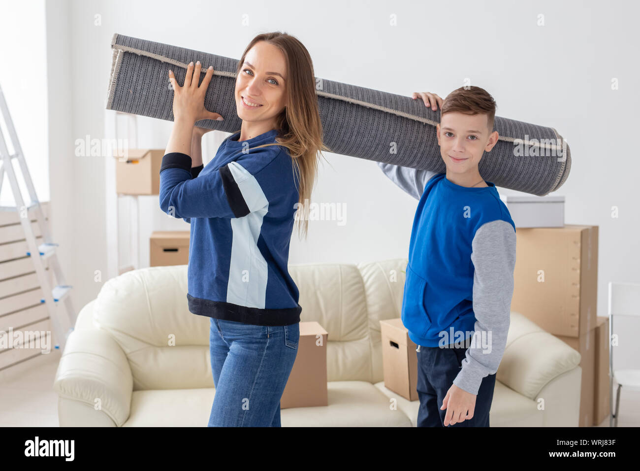 Smiling caucasian single mother and a charming son are holding a folded carpet on their shoulders in a new living room intending to spread it in a new Stock Photo