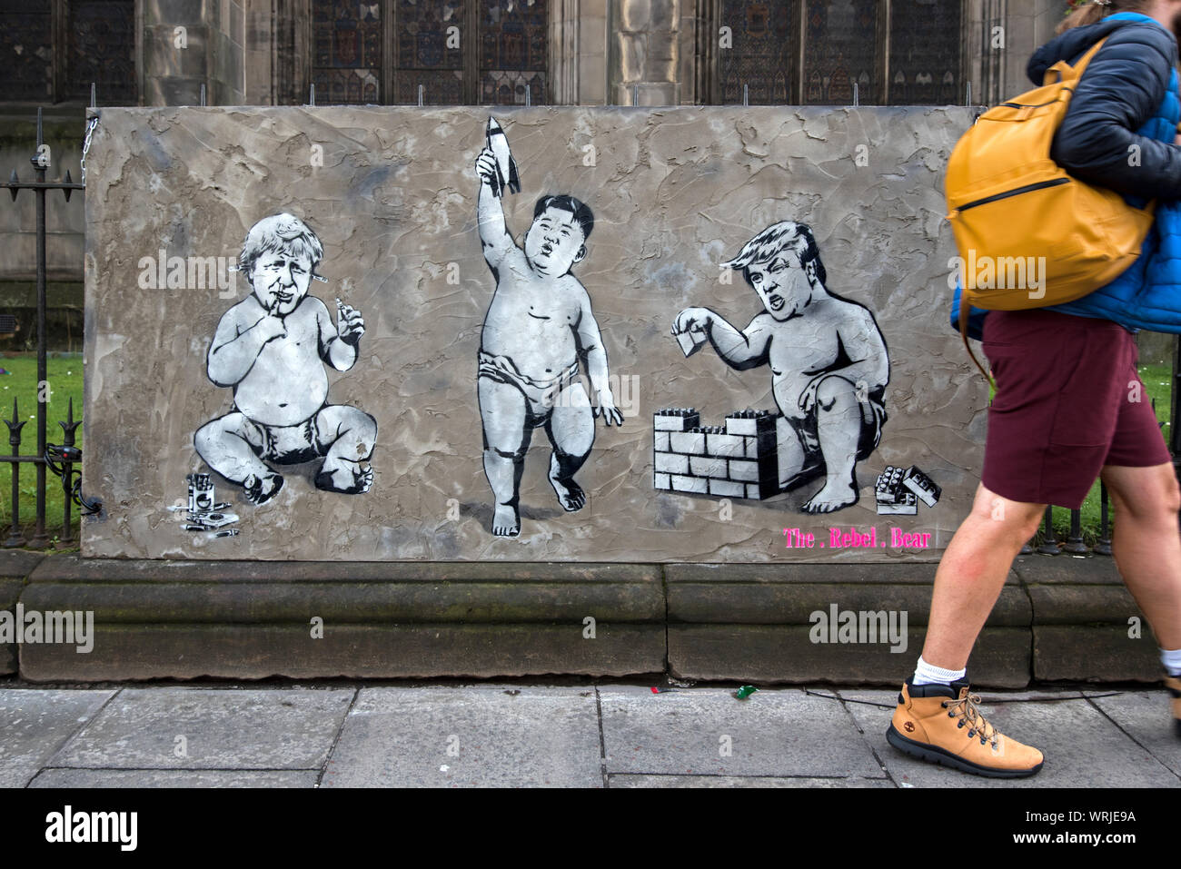 Pedestrian walking by piece of street art depicting Donald Trump, Boris Johnson and Kim Jong Un as babies playing with toys. Edinburgh, Scotland. Stock Photo