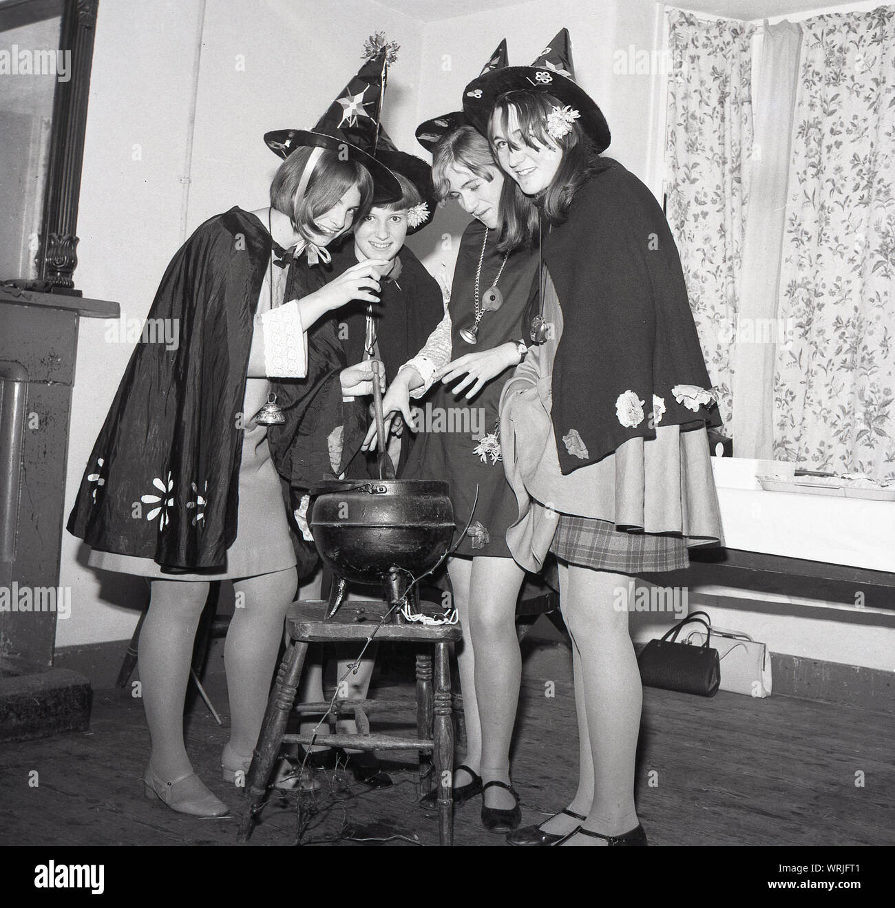 1967, historical, four teenage girls dressed up in witches costumes - black  coats and pointed hats - standing in a room around a cast iron pot or  cauldron perched on a stool,