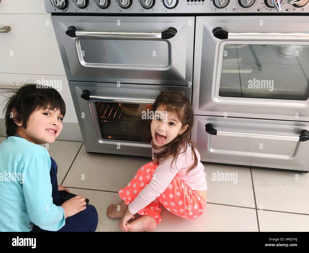 Portrait Of Siblings By Oven On Floor In Kitchen At Home Stock Photo