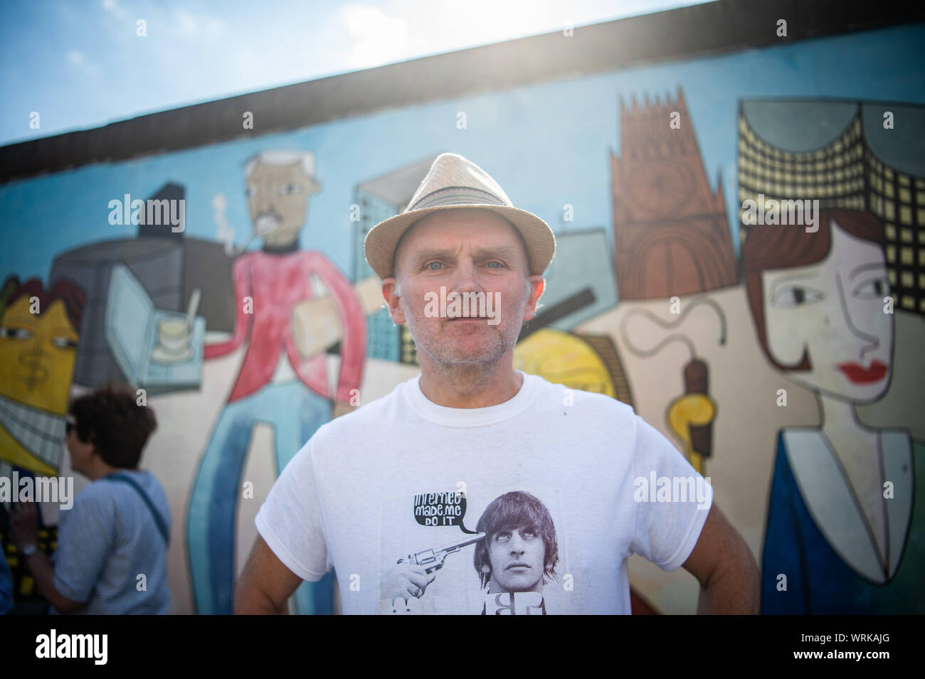 22 August 2019, Berlin: Jim Avignon, artist, stands in front of his work at the East Side Gallery. Almost 30 years ago, artists immortalized themselves here and reinterpreted the longest remaining section of the Berlin Wall with their works of art. Photo: Arne Immanuel Bänsch/dpa Stock Photo