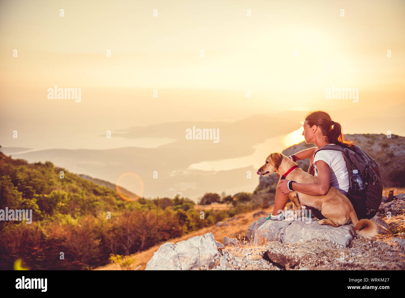 Woman with a dog sitting on a mountain and looking at a bay during sunset Stock Photo