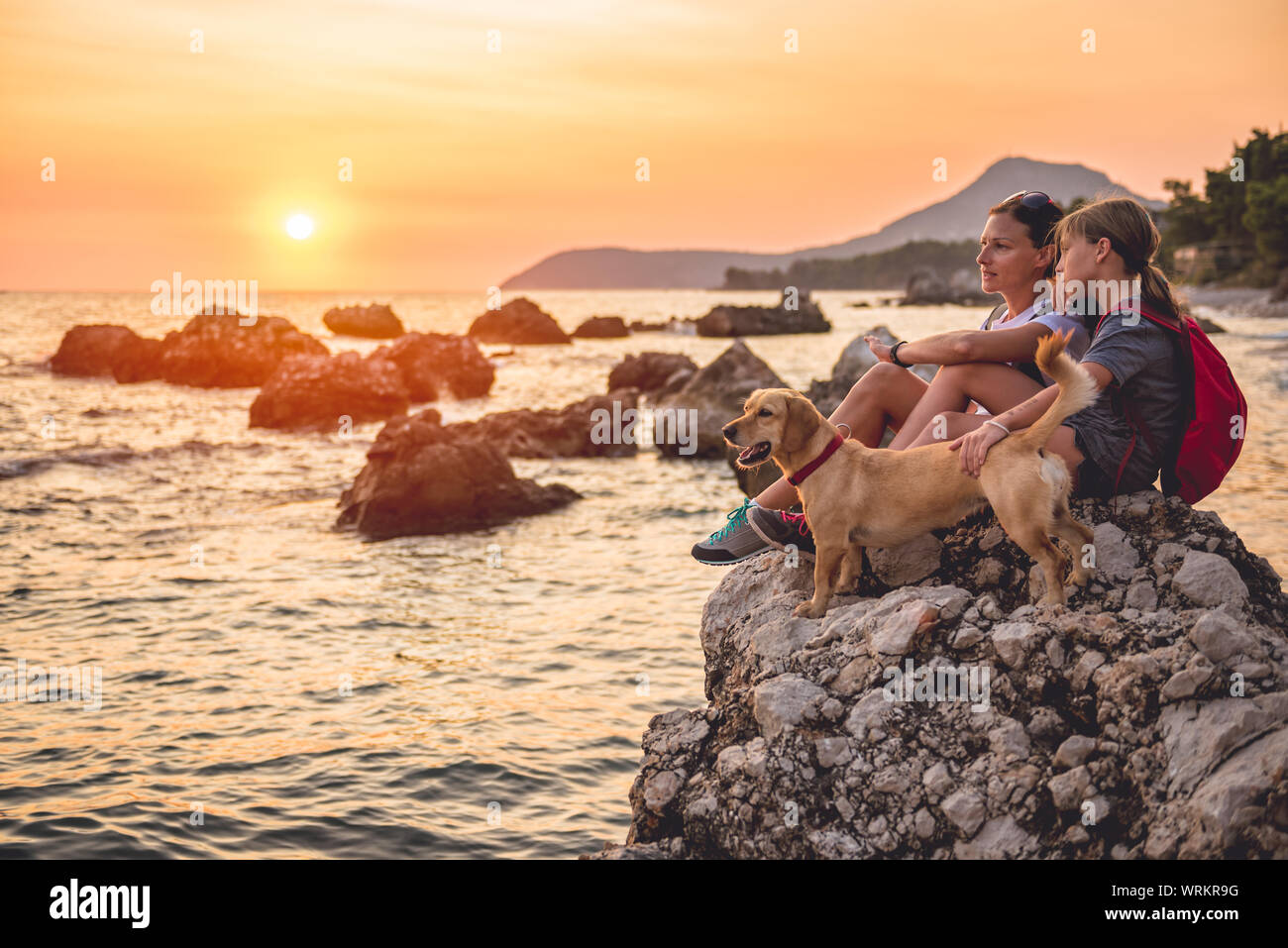 Mother and daughter with a small yellow dog hiking along the seashore during sunset Stock Photo