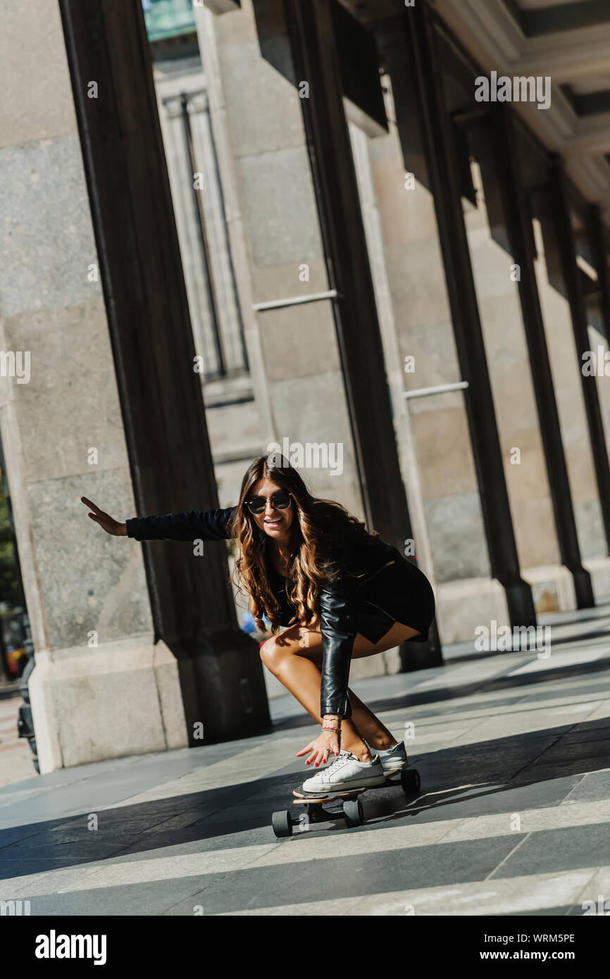 Beautiful young skater woman riding on her longboard in the city. Stylish girl in street clothes rides on a longboard. Stock Photo