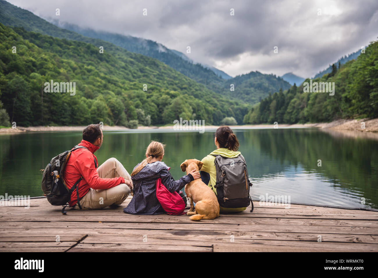 Family with a small yellow dog resting on a pier and looking at lake and foggy mountains Stock Photo