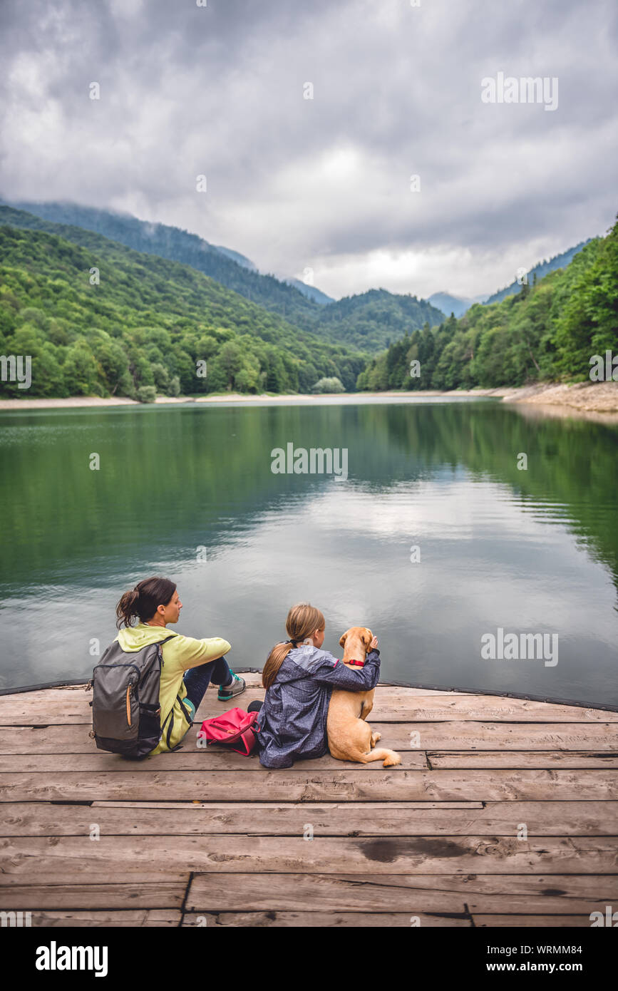 Mother and daughter with a small yellow dog resting on a pier and looking at lake and foggy mountains Stock Photo