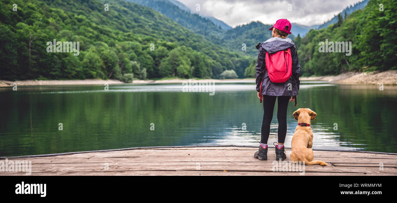 Girl with a small yellow dog standing on pier by the mountain lake Stock Photo