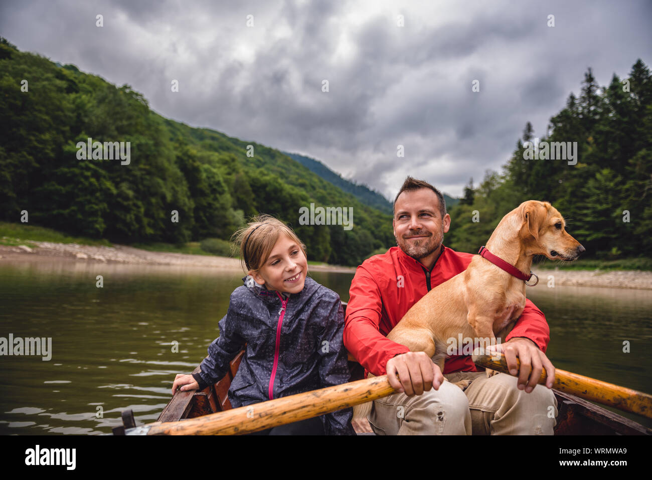 Father and daughter with a small yellow dog rowing a boat on a mountain ...
