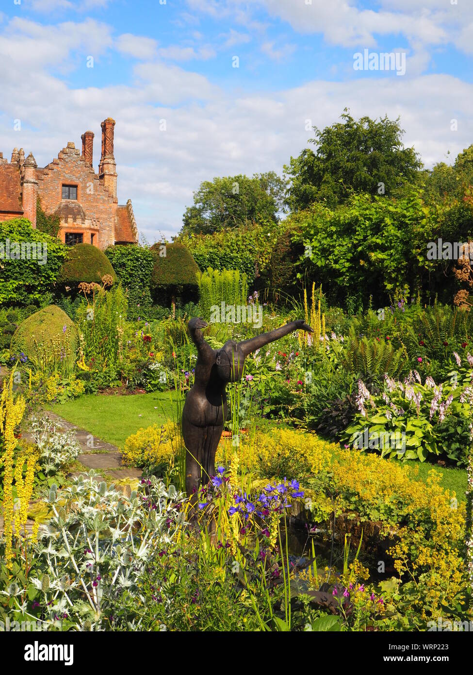 Portrait of Chenies Manor house and sunken garden, ornamental pond with Alan Biggs' sculpture;f resh green plant border in late afternoon sun in June. Stock Photo