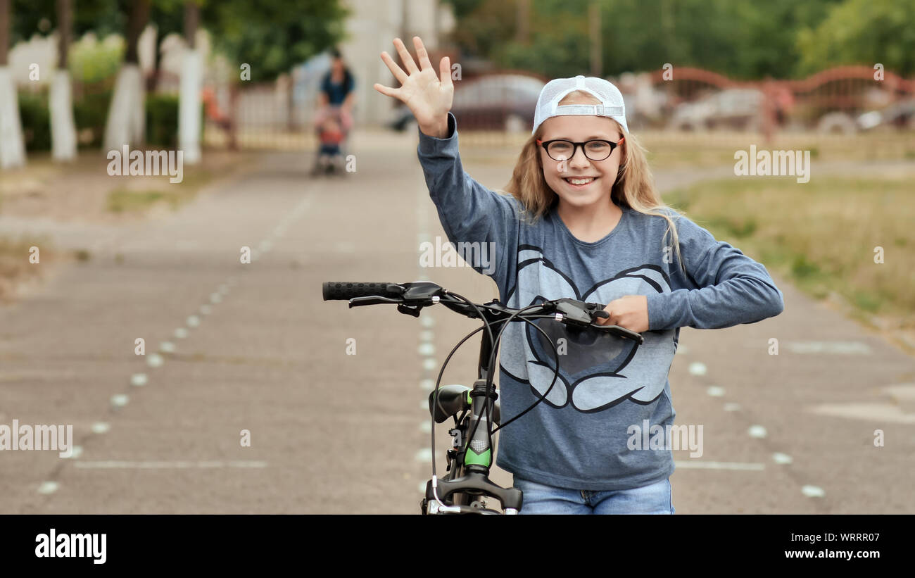 An eleven-year-old girl in a good mood rides a bicycle and after a stop shows a thumbs-up. Stock Photo
