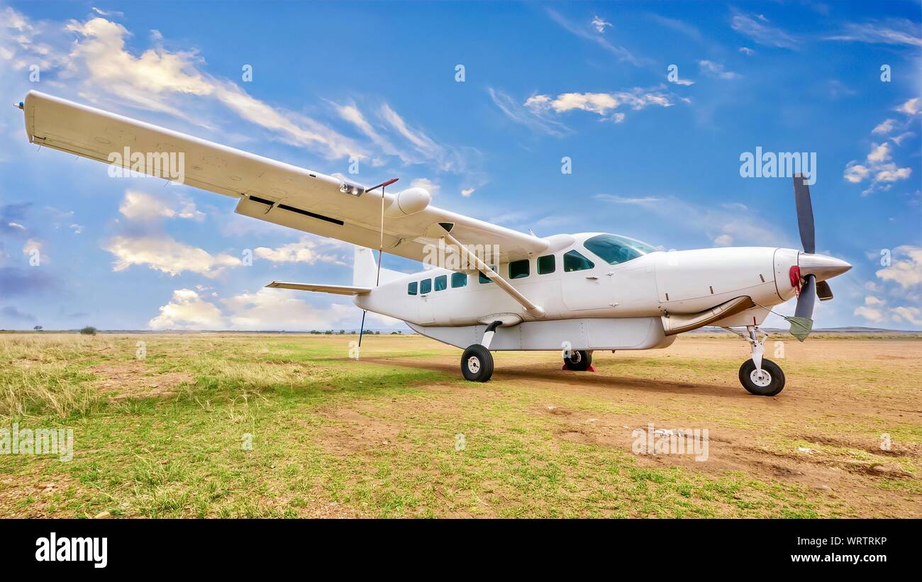 A white, single engine charter plane sits on a grass and dirt landing strip in a beautiful and remote location in Kenya Stock Photo