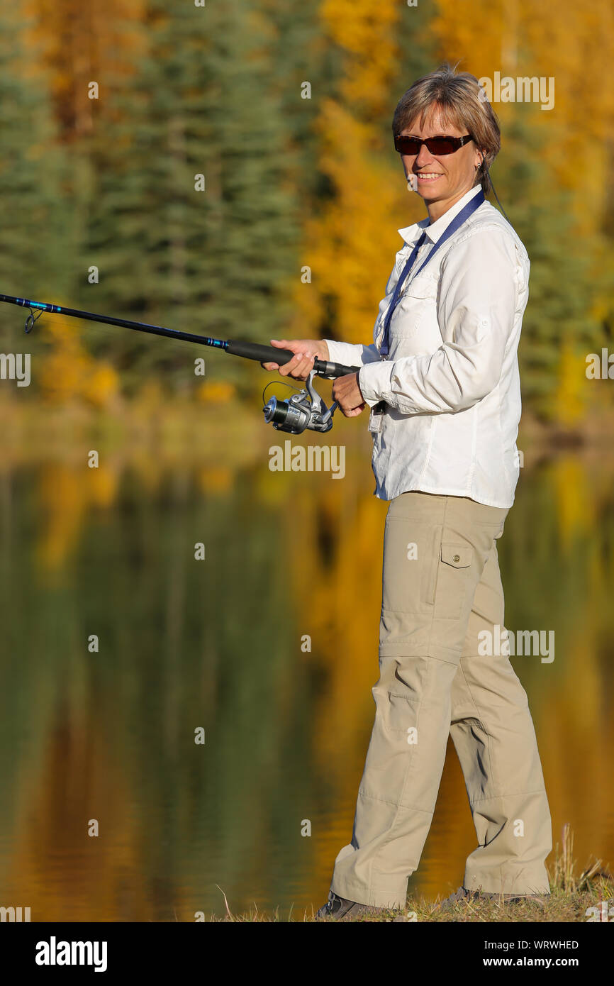 Woman standing at a lake with a rod, facing camera, colorful trees and reflection in lake Stock Photo