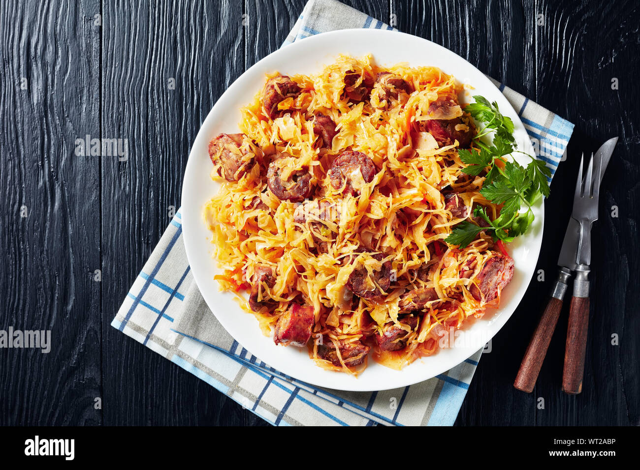 delicious Cabbage stew with sliced fried sausages served on a white plate on a black wooden table, view from above, flat lay, close-up Stock Photo