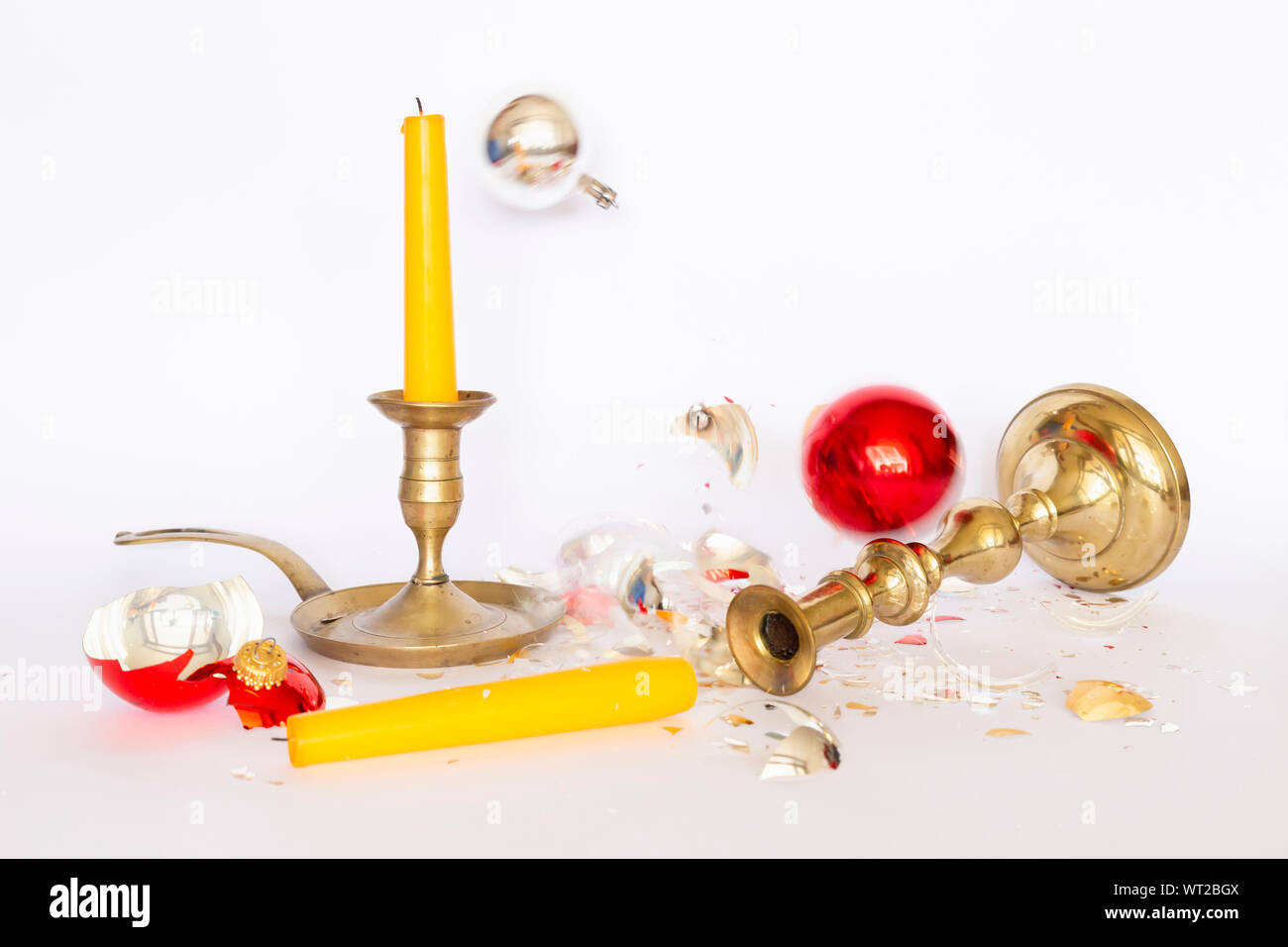 Front view of falling and shattered red and silver Christmas baubles and two bronze candleholders with a yellow candle on white background Stock Photo