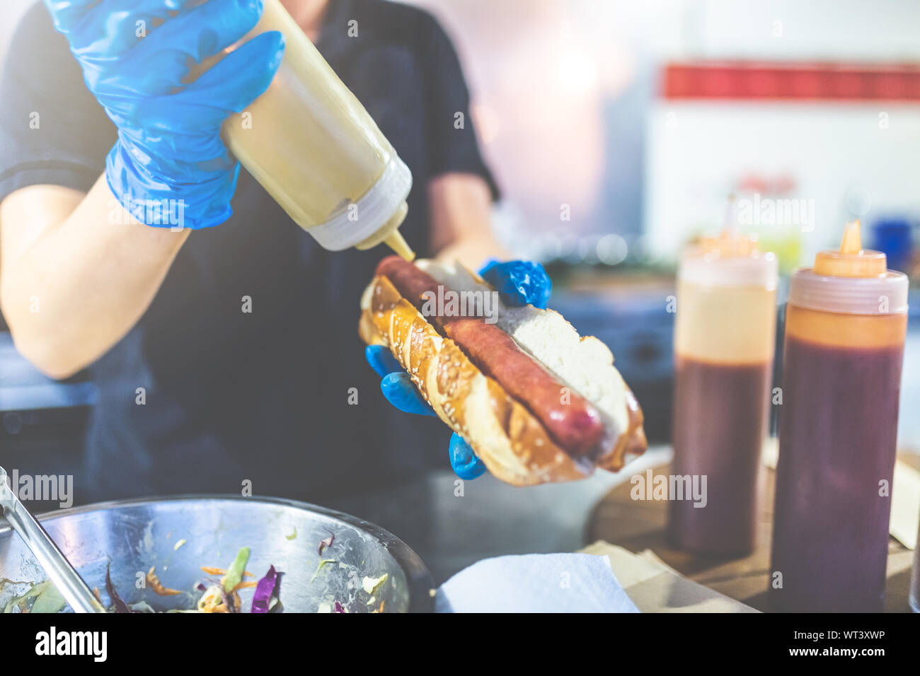 Close up of chef hands pouring mustard on fresh hot dog with grilled sausage. Stock Photo