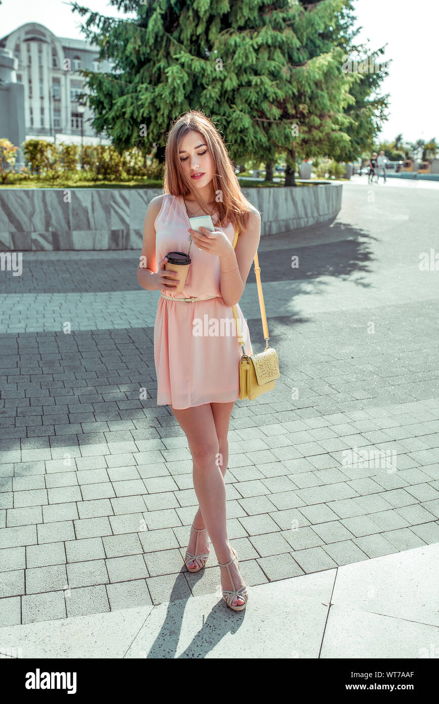 Girl in summer city, pink dress sandals. In hands of phone is an on-line  application personal networks, cup coffee tea. Handbag over shoulder, view  Stock Photo - Alamy