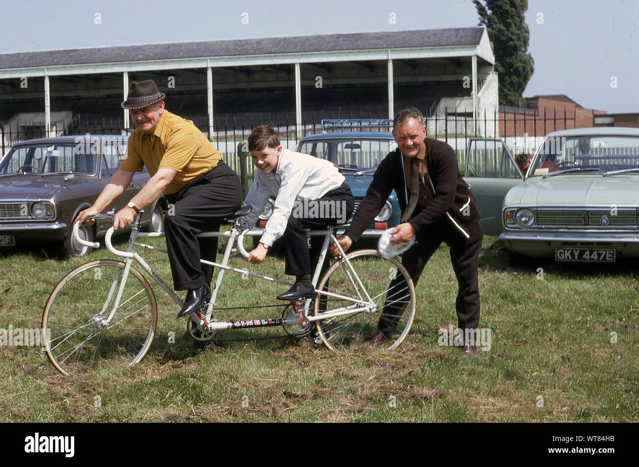 1960s, historical, Outside at the annual CTC rally at York racecourse, a man and a boy sitting on a 'Cincelli' tandem bicycle. Cars of the era, including Ford Cortinas are parked in the field. Stock Photo