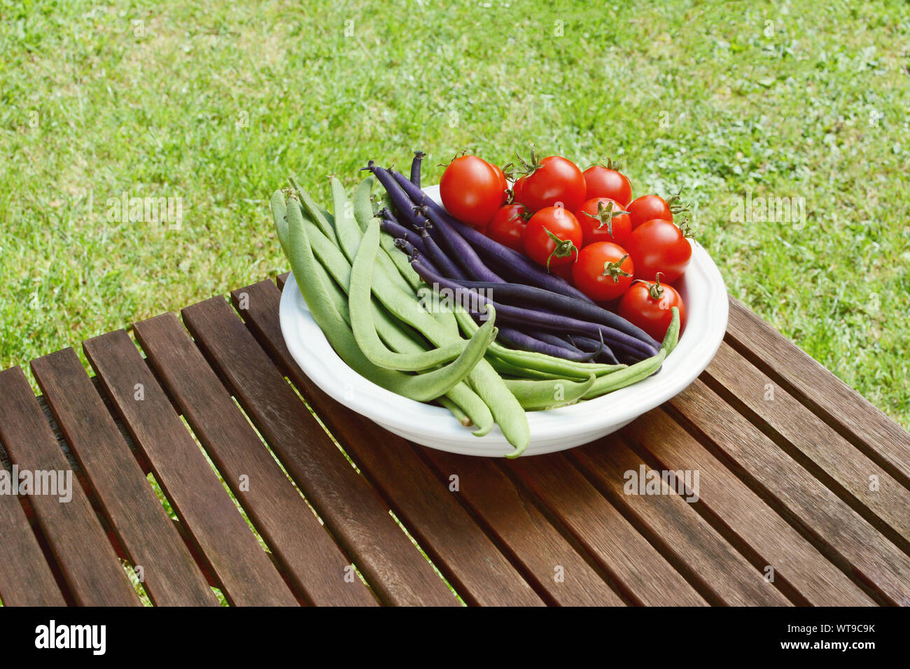 Freshly harvested Calypso beans, French beans and cherry tomatoes in a dish on a wooden picnic table in a garden Stock Photo