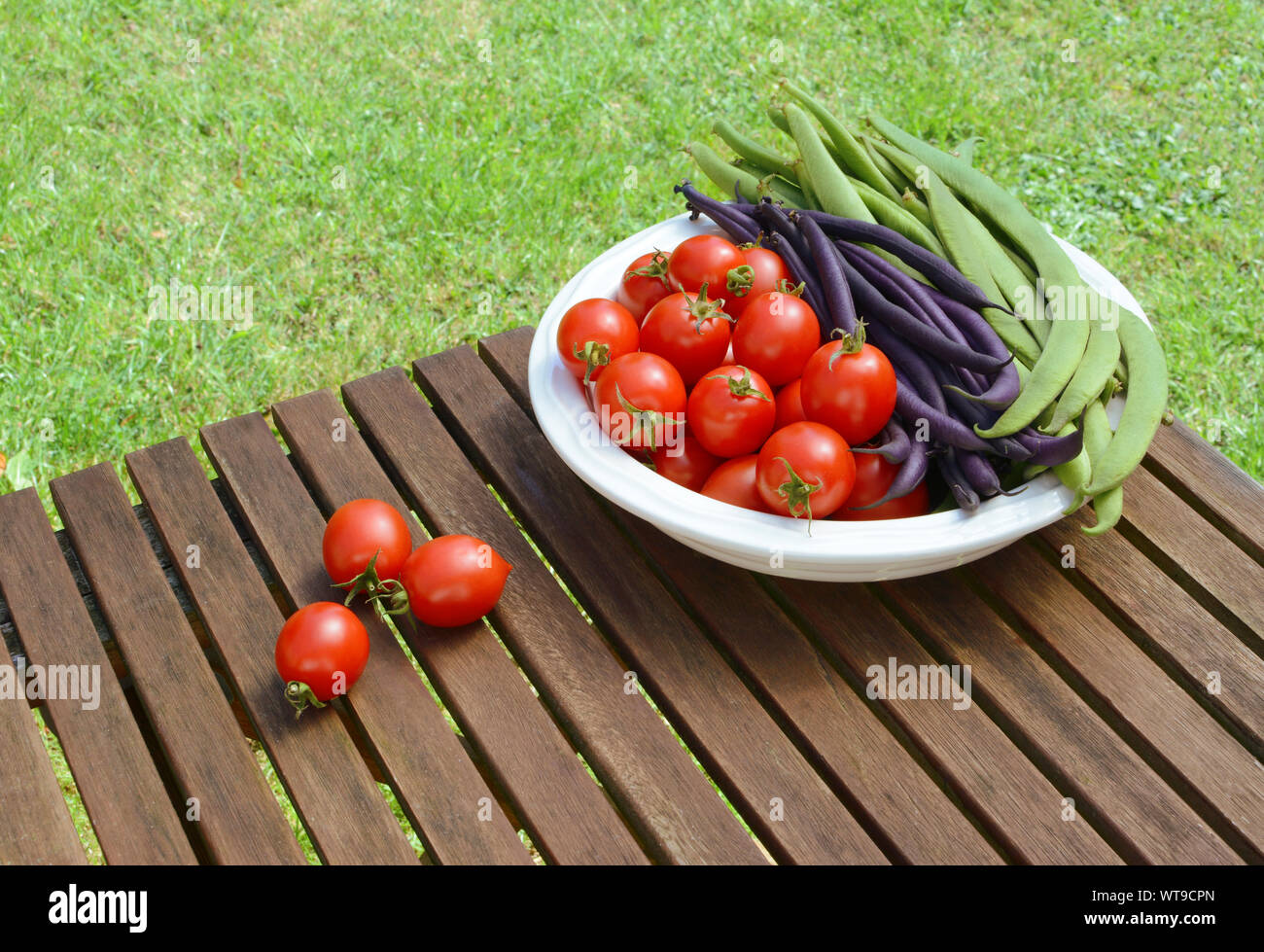 Red tomatoes with green and purple beans in a dish, some cherry tomatoes spilled on a wooden picnic table Stock Photo
